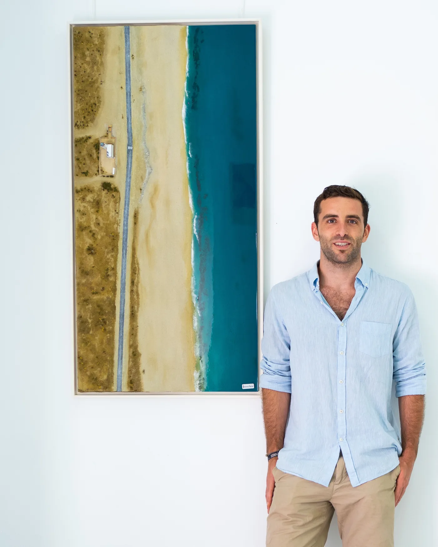 Álvaro Varela de pie junto a una obra vertical enmarcada: una playa larga con carretera, dunas y mar azul.