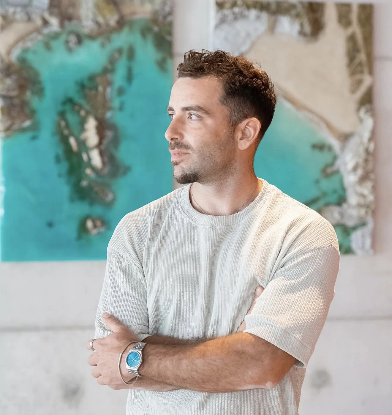 Retrato de Álvaro Varela, de brazos cruzados, delante de dos de sus obras de mar turquesa y roca.
