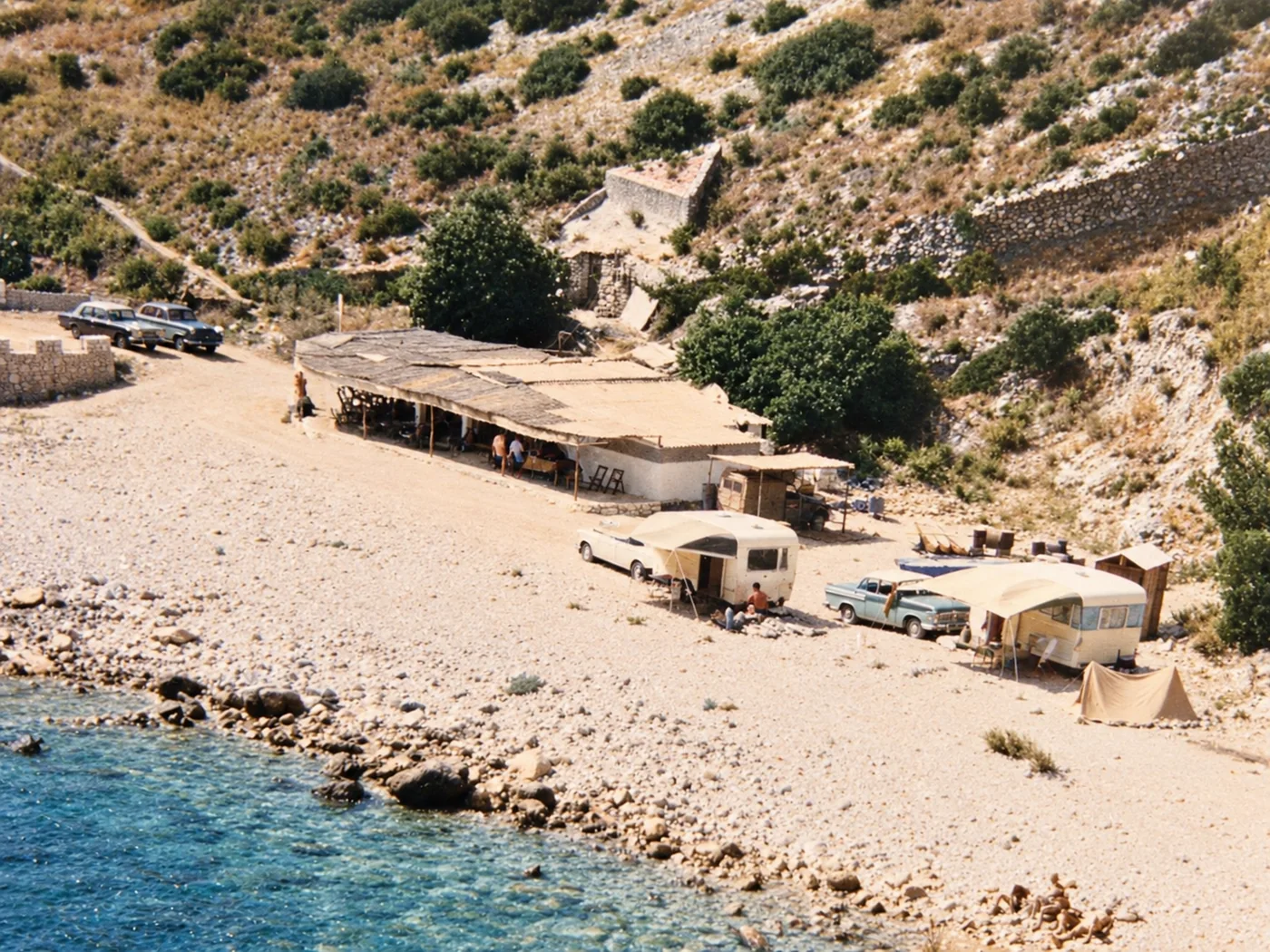 Fotografía antigua: la playa de cantos con un chiringuito de cañizo, coches de época, caravanas y una tienda de campaña.
