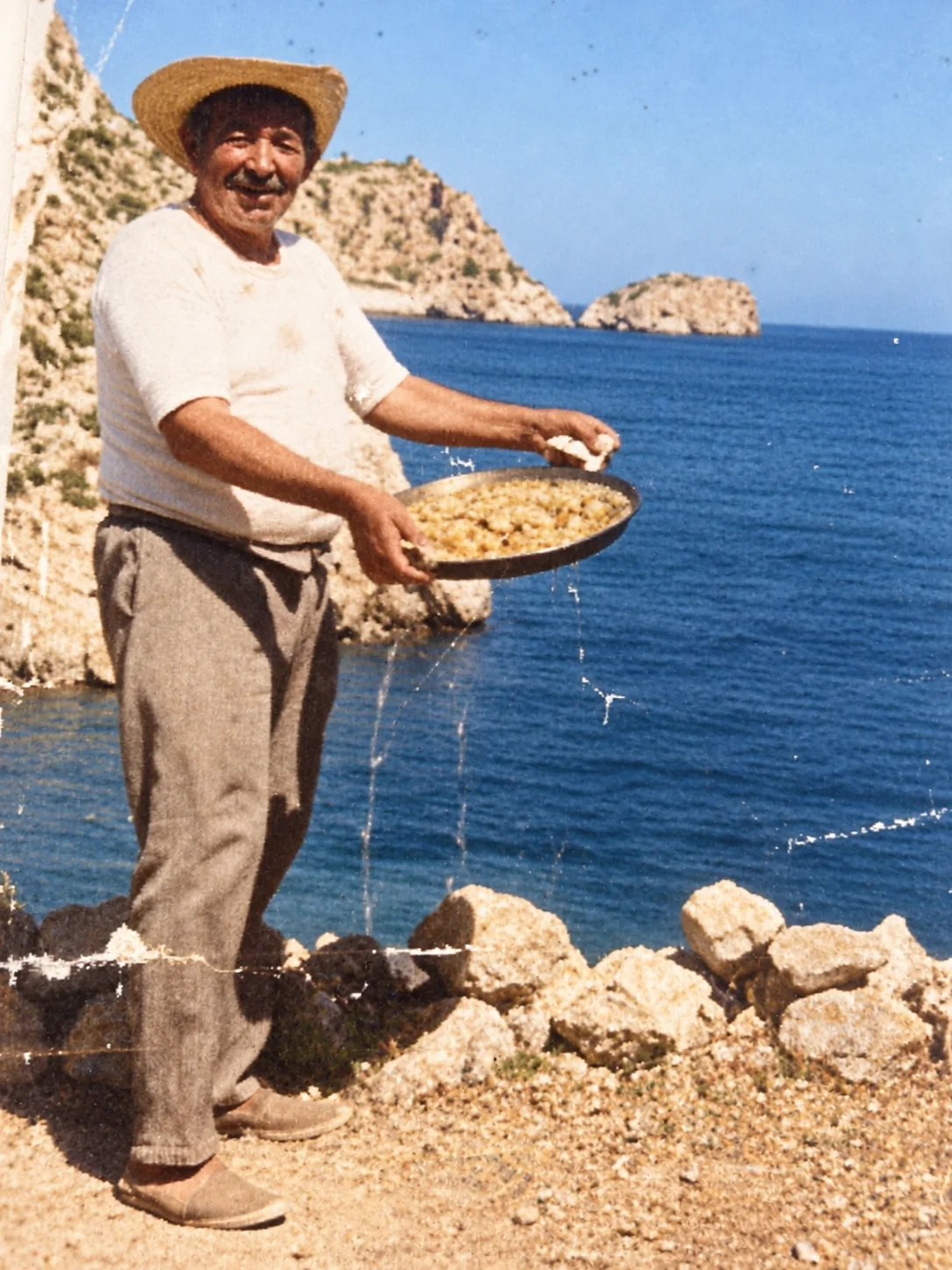 Fotografía antigua en color: un hombre con sombrero de paja y alpargatas sonríe y sostiene una paella junto al mar, con un islote al fondo.