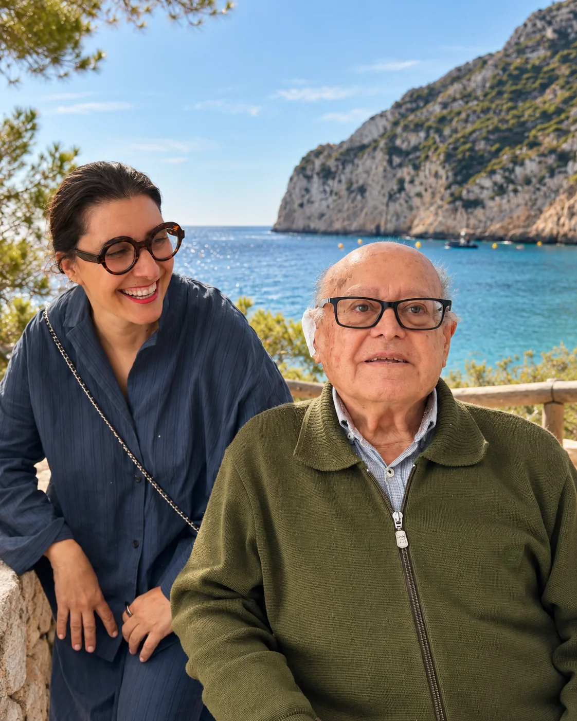Don Salvador Ginestar Soler, seated, smiling at the camera. Beside him, Jessica Bataille looks at him, laughing. Behind them, the sea and the cliff of La Granadella.